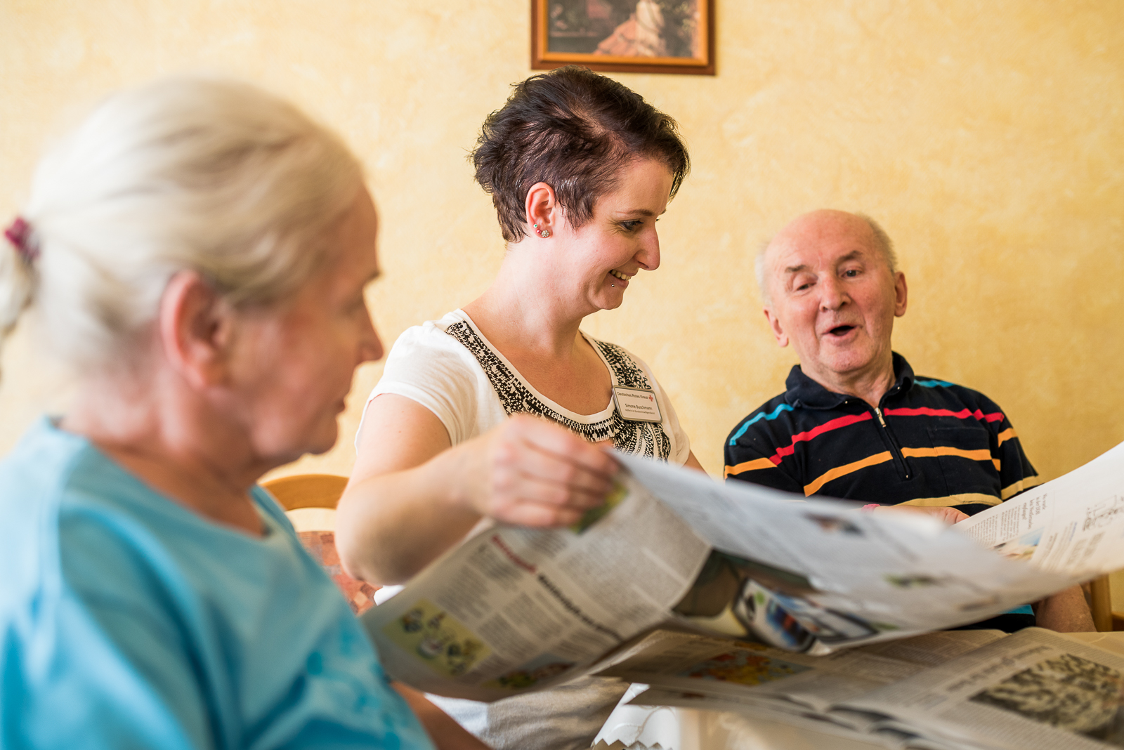 Eine Frau mittleren Alters sitzt mit zwei Senioren, einer Frau und einem Mann, an einem runden Tisch. Sie liest den beiden aus der Zeitung vor und der ältere Mann lacht über etwas.