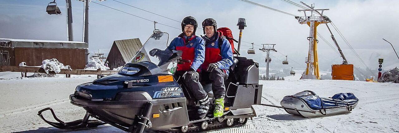 Zwei Bergretter sitzen auf einem Schneefahrzeug neben einem Skilift. Sie sichern die Skipiste ab.