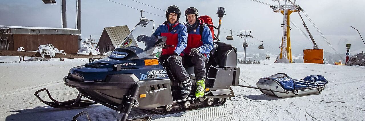 Zwei Bergretter sitzen auf einem Schneefahrzeug neben einem Skilift. Sie sichern die Skipiste ab.