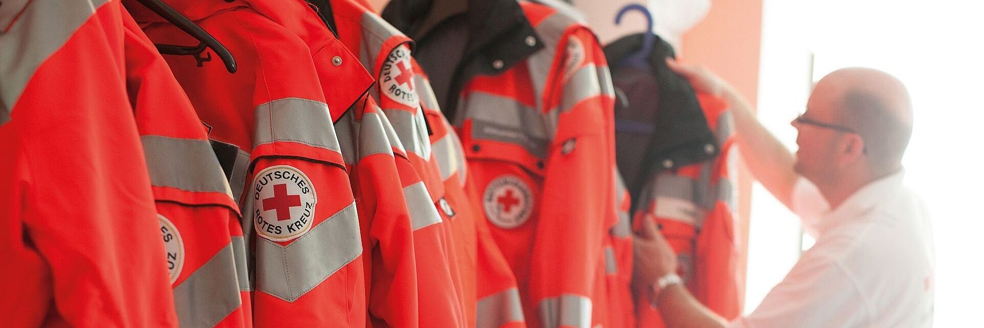 Ein Mitglied des DRK hängt seine Rettungsjacke auf einen Haken. Daneben hängen einige weitere DRK-Rettungsjacken in leuchtendem Orange.