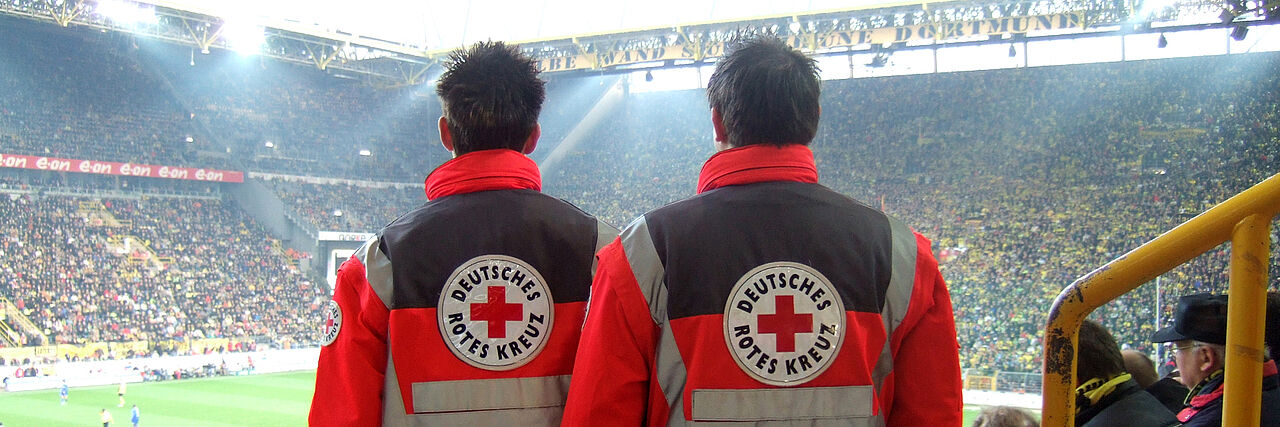 Zwei Rettungssanitäter stehen während eines Fußballspiels auf dem Rang im Stadion und sind bereit für den Einsatz im Notfall.